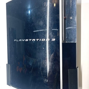 Peut inclure: Une console PlayStation 3 noire avec une plaque frontale argentée et le texte "PLAYSTATION 3" imprimé dessus. La console est fixée au mur avec un support noir.