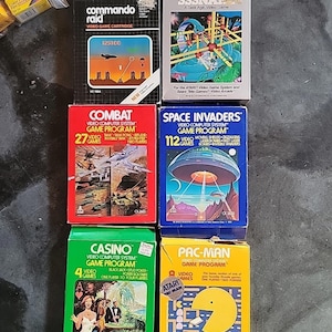 Puede incluir: Una colección de cartuchos de videojuegos Atari vintage, incluyendo "Commando Raid", "Combat", "Space Invaders", "Casino" y "Pac-Man". Las cajas presentan gráficos y texto coloridos, mostrando los títulos y características de los juegos.