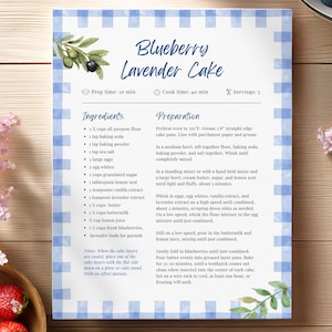May include: A recipe card for Blueberry Lavender Cake with a blue gingham border. The card includes ingredients and preparation steps. Fresh strawberries and blueberries are in a wooden bowl and a white plate. Pink flowers are on the left and right.