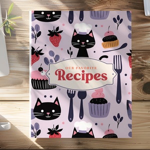 Könnte beinhalten: Ein rosa-weißer Rezeptbuchumschlag mit einer schwarzen Katze, Cupcakes und Erdbeeren. Der Text auf dem Umschlag lautet "Unsere Lieblingsrezepte".