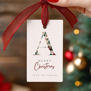 May include: A white gift tag with a red ribbon, featuring a floral-patterned letter "A". The tag reads "Merry Christmas" and "From The Abrams" in a burgundy script. The background includes Christmas ornaments.