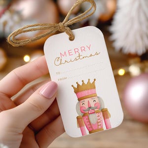 May include: A white gift tag with a watercolor illustration of a pink and gold nutcracker. The tag has the words "MERRY Christmas" in pink script, with space to write "TO" and "FROM". A natural twine bow is at the top.