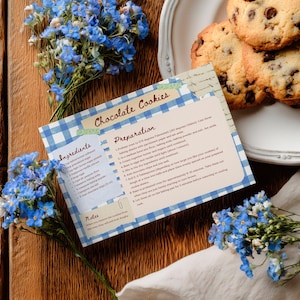 May include: A photograph of a recipe card for chocolate cookies, a plate of cookies, and blue flowers. The recipe card has a blue and white checked border and the words "Chocolate Cookies". The cookies are golden brown with chocolate chips.