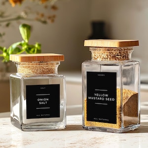 Peut inclure: Deux pots à épices en verre avec des couvercles en bois. Le pot de gauche contient du sel d'oignon et le pot de droite contient des graines de moutarde jaune. Les deux pots ont des étiquettes noires avec du texte blanc.