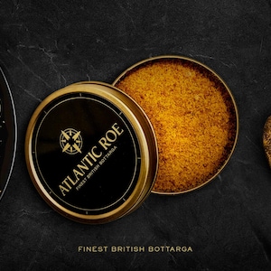 May include: A top-down shot features a black plate of spaghetti, a wooden spoon, and a tin of Atlantic Roe Bottarga. The tin is open, revealing the roe. The text reads "Finest British Bottarga."