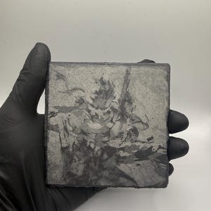May include: A square slate coaster featuring a dark, monochromatic ink-style illustration of a character holding a sword. The coaster has a rough, natural stone texture with jagged edges and a grey, weathered appearance throughout the surface.