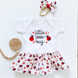 May include: White baby onesie with a red ladybug print and the text "little love bug". The onesie has a white ruffle skirt with a red ladybug print. A red ladybug headband is included.
