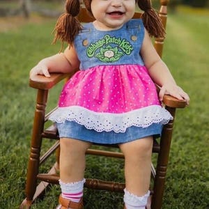 May include: A brown yarn wig with pigtails, a blue denim jumper dress with a Cabbage Patch Kids logo, a pink polka dot skirt section, a white eyelet lace trim, white ruffled socks, and brown leather-style shoes with white accents.