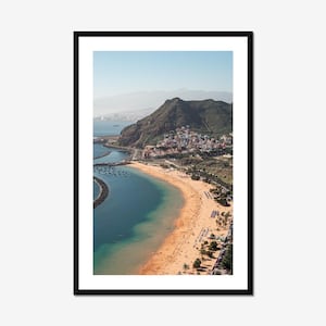 Puede incluir: Vista aérea de una playa con arena dorada y agua turquesa, enmarcada por un borde negro. Una montaña con exuberante vegetación verde se alza en el fondo, con un pueblo en sus laderas. El cielo es azul claro.