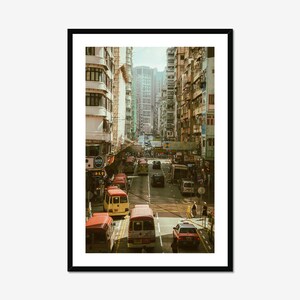 Puede incluir: Fotografía enmarcada de una concurrida escena callejera en una ciudad. La imagen presenta una carretera llena de vehículos, incluyendo autobuses y coches, flanqueada por edificios altos. La paleta de colores es cálida, con una estética vintage.
