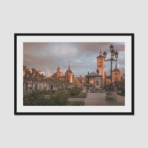 May include: A framed photograph of a European city square with buildings, trees, and a statue. The sky is overcast with a rainbow visible. The photograph is in a black frame.