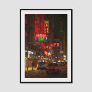 Puede incluir: Una impresión de arte mural vertical enmarcada que representa una escena nocturna de una calle en Hong Kong. La imagen muestra un taxi rojo circulando por una carretera bordeada de edificios altos cubiertos de letreros de neón rojo y verde.