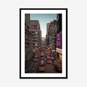 Puede incluir: Fotografía enmarcada de una concurrida calle de Hong Kong. La imagen muestra autobuses rojos y blancos, coches y peatones. Los edificios bordean la calle y se ven letreros de neón. El cielo es de un azul suave.