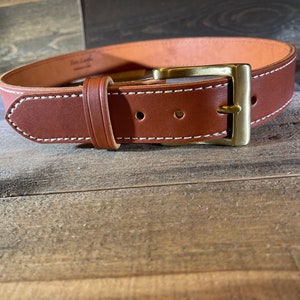 May include: A brown leather belt with white stitching and a brass buckle. The belt is on a wooden surface.