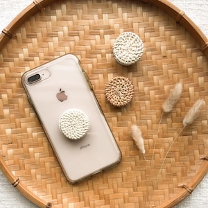 May include: A white wicker phone grip attached to a clear phone case with the word "iPhone" printed on the back. The phone case is on a woven tray with two other white wicker phone grips.