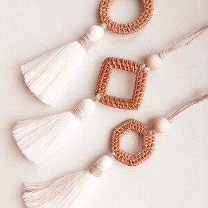 Może przedstawiać: Trzy białe breloki z frędzlami i różnymi kształtami z plecionego drewna. Kształty to koło, kwadrat i sześciokąt.