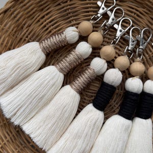 Może przedstawiać: Zestaw breloków z białymi frędzlami, drewnianymi koralikami i srebrnymi zapięciami. Niektóre frędzle mają brązowe akcenty z nici, inne czarne. Breloki ułożone są na wiklinowym koszu.