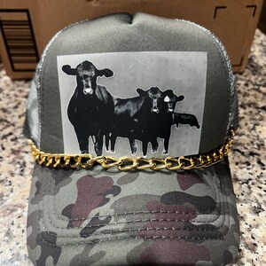 May include: A green camouflage trucker hat with a gold chain trim. The hat features a black and white image of two cows on the front.