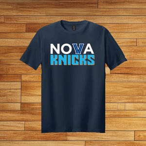 May include: Navy blue t-shirt with the words "NOVA KNICKS" in white and blue lettering. The shirt has a crew neck and short sleeves. The background is a wooden surface.