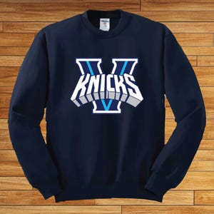 May include: A navy blue long-sleeve crewneck sweatshirt featuring a large graphic on the chest. The design displays a stylized letter V in light blue and white, with the word KNICKS written in bold white block letters across the center.