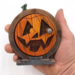May include: A wooden Halloween decoration shaped like a jack-o'-lantern door. The door is orange with a carved face and a silver knob. The frame is brown, and the base is grey. A green stem sits on top.