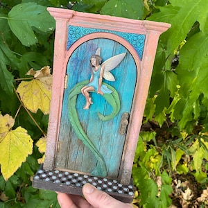 May include: A fairy door with a teal door and a pink frame. A fairy in a blue dress sits on a green leaf. The door has a decorative handle and a small step with a dotted pattern.