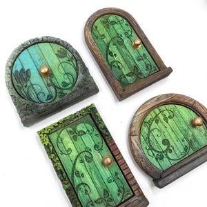 Puede incluir: Cuatro puertas de hadas en miniatura de varias formas. Cada puerta presenta un diseño de madera con una combinación de colores verde y azul, adornada con patrones florales y un pomo dorado. Las puertas están sobre un fondo blanco.