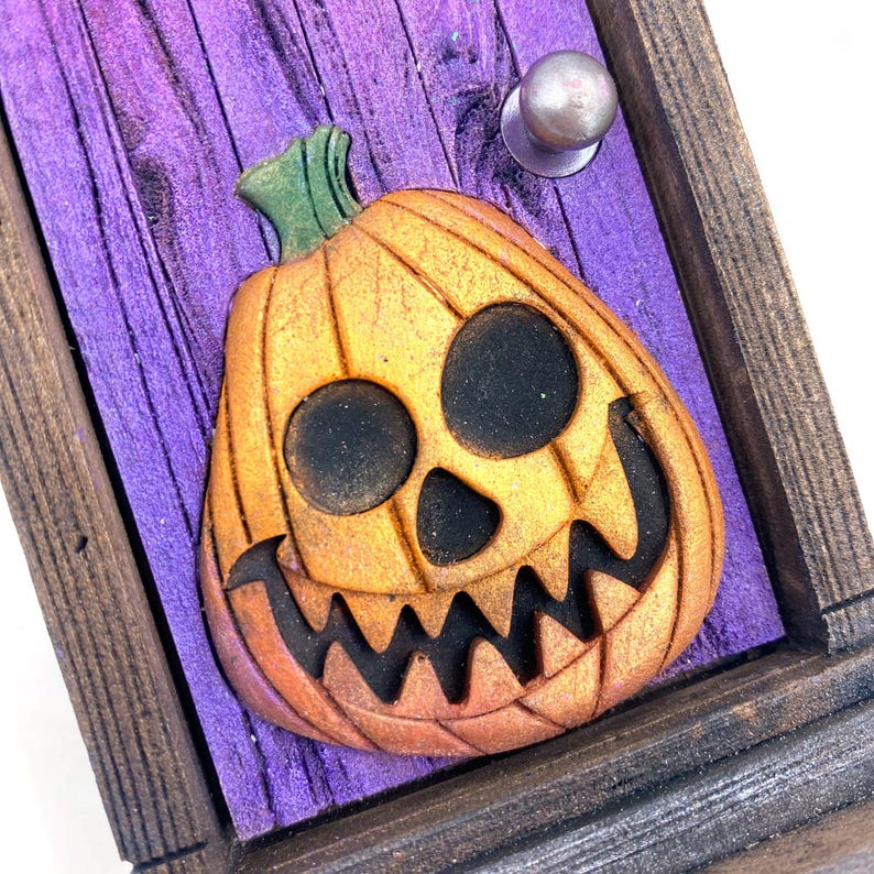 May include: A Halloween-themed decorative wooden plaque featuring a carved orange pumpkin with a jack-o'-lantern face. The pumpkin is set against a purple wooden background with a silver knob and framed by a dark brown wooden border.