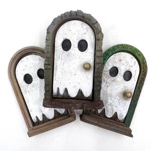 May include: Three decorative ghost-shaped doors. Each has a white ghost face with black eyes and a brass doorknob. The frames are in shades of brown and green, creating a spooky Halloween theme for seasonal decor.