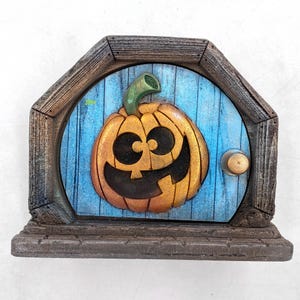 May include: A decorative pumpkin-themed fairy door with a smiling jack-o'-lantern face. The door is painted blue with a wooden frame and a small brass doorknob. The pumpkin is orange with a green stem.