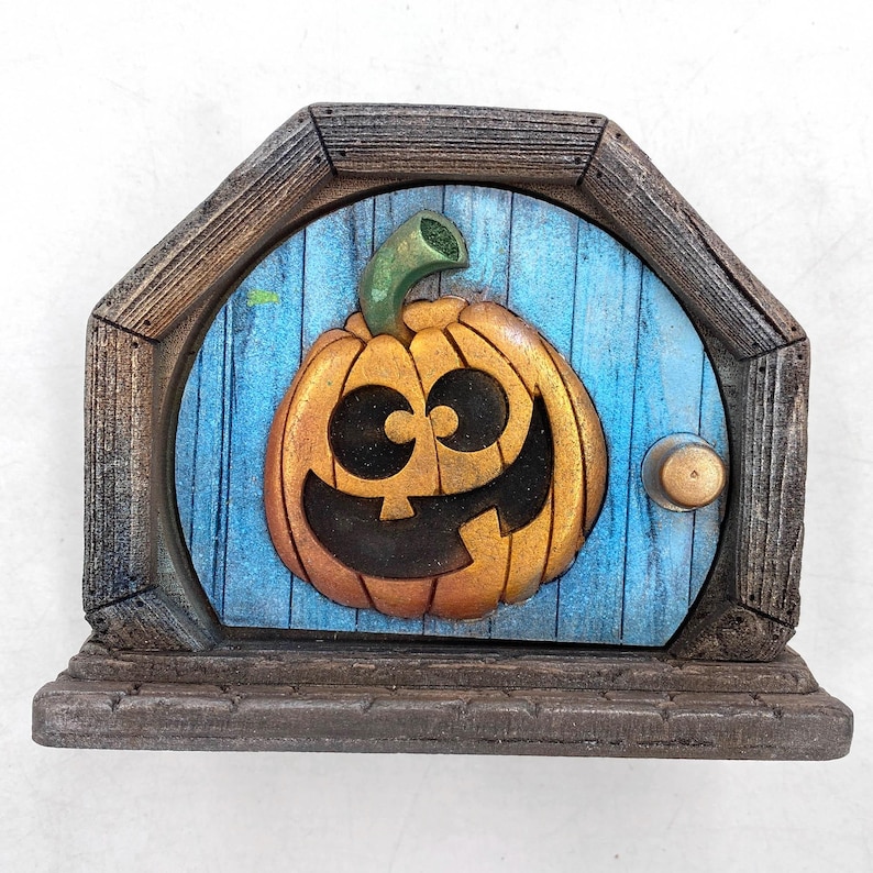May include: A decorative pumpkin-themed fairy door with a smiling jack-o'-lantern face. The door is painted blue with a wooden frame and a small brass doorknob. The pumpkin is orange with a green stem.