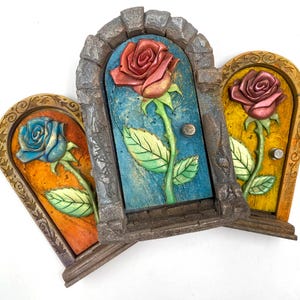 May include: Three decorative rose sculptures shaped like arched doors. Each door features a different coloured rose: blue, red, and pink. The doors have a textured appearance with a stone-like frame and a painted background.