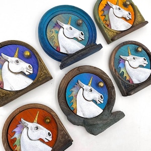 May include: Six decorative wall plaques, each depicting a white unicorn head with a golden horn. The plaques have circular frames in various colours, including blue, brown, and green. A small, gold sphere is positioned above the unicorn's head. The background colours vary.