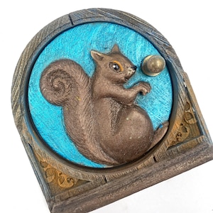 May include: A brown squirrel sculpture with a golden acorn, set against a turquoise background. The squirrel is in a detailed relief, framed by a wooden-look border. A decorative piece for home decor.