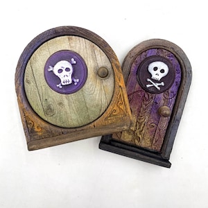 Puede incluir: Dos puertas decorativas de madera con un emblema de calavera y huesos cruzados. Una puerta es redonda con un acabado verde y marrón, la otra es arqueada con un acabado morado y marrón. Ambas tienen un pequeño pomo, probablemente para un jardín de hadas.