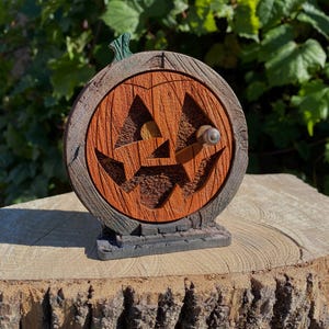 May include: A decorative Halloween pumpkin door with a carved jack-o'-lantern face. The door is orange with a wooden texture, set in a dark brown frame. A small silver knob is on the door. The base is a dark gray stone.