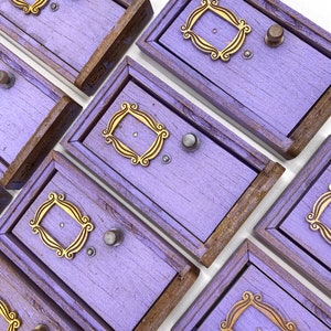 May include: Several small, lavender-painted wooden boxes with a distressed finish. Each box has a gold-coloured decorative frame and a silver knob. The boxes are arranged diagonally, highlighting their unique design and rustic charm.