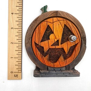 May include: A wooden Halloween decoration shaped like a pumpkin door. The door is orange with a carved jack-o'-lantern face and a silver doorknob. The door is set in a dark brown wooden frame and sits on a gray stone base. The door is approximately 4 inches tall.