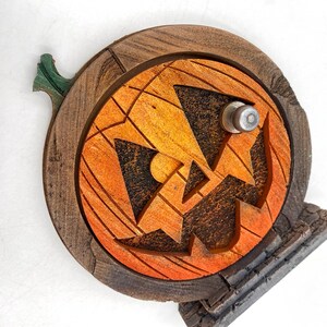 May include: A wooden Halloween decoration featuring a carved orange pumpkin face with a jack-o'-lantern design. The pumpkin is set within a dark brown wooden frame and includes a green stem. The base is a gray stone-like structure.