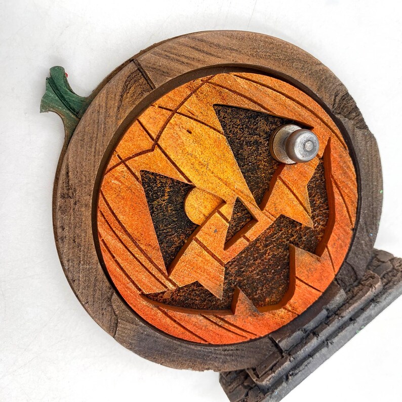 May include: A wooden Halloween decoration featuring a carved orange pumpkin face with a jack-o'-lantern design. The pumpkin is set within a dark brown wooden frame and includes a green stem. The base is a gray stone-like structure.
