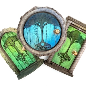 Puede incluir: Tres puertas de hadas en miniatura en tonos verdes y azules. Cada puerta presenta un diseño de árbol y un pomo de color latón. Las puertas están enmarcadas con texturas similares a la madera, creando una estética caprichosa y de fantasía.