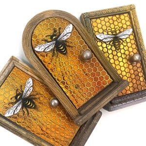 May include: Three decorative wooden boxes with a bee theme. Each box features a honeycomb pattern background, a detailed bee, and a small silver knob. The boxes have a rustic, aged appearance with a mix of brown, gold, and silver tones.