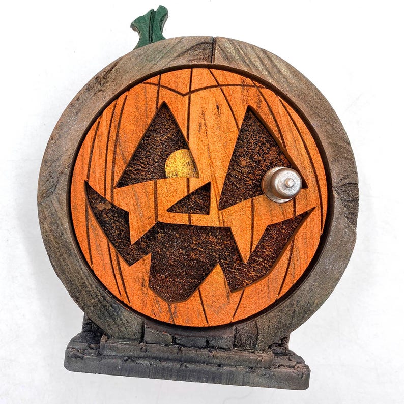 May include: A wooden Halloween decoration shaped like a pumpkin door. The pumpkin has an orange face with carved-out eyes and a mouth, a silver doorknob, and a green stem. The door is set in a weathered wooden frame and sits on a gray stone base.
