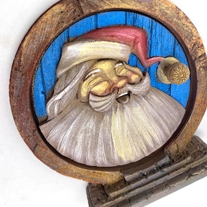 May include: A wooden, circular Christmas ornament featuring a carved Santa Claus face. The Santa has a red hat, white beard, and gold accents. The background is blue with a wooden frame and a decorative base.