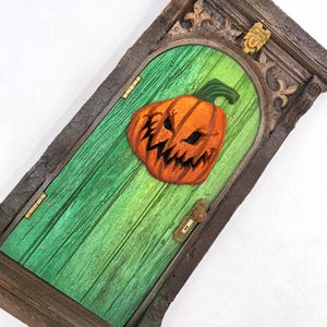 Puede incluir: Una puerta en miniatura con temática de Halloween con una cara de calabaza naranja tallada. La puerta verde tiene una textura de vetas de madera y está colocada en un marco de piedra. Se ve una pequeña manija y bisagras doradas.