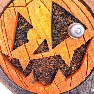 May include: Close-up of a wooden jack-o'-lantern face with a carved, toothy grin and triangular eyes. The pumpkin is painted orange with black accents in the carved areas and a silver metal eye bolt.