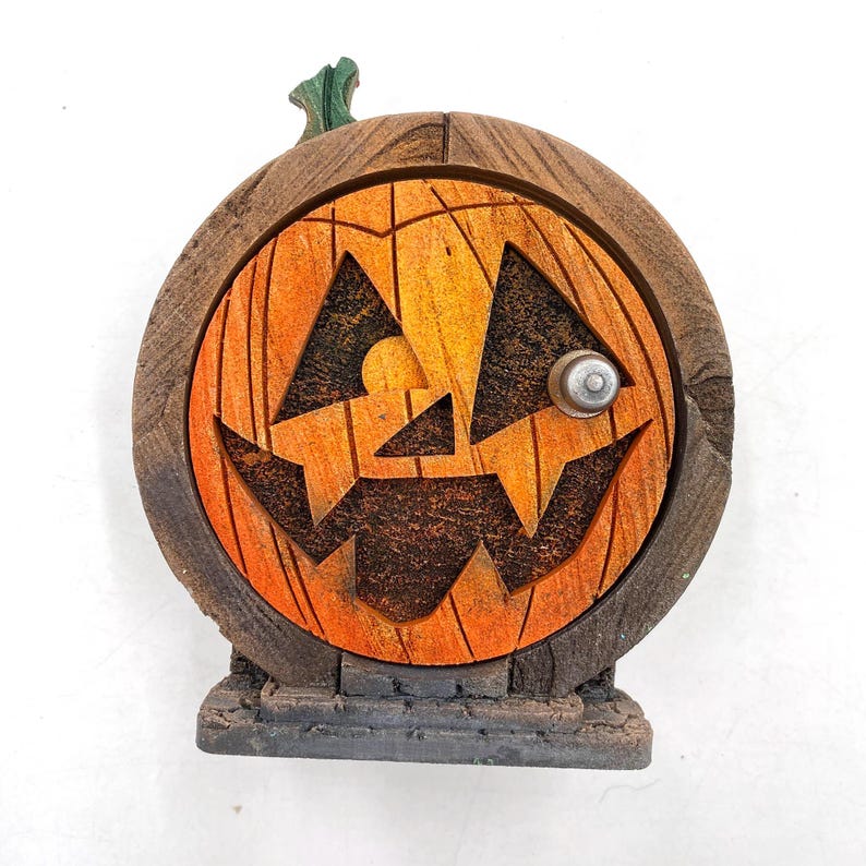 May include: A Halloween-themed decorative pumpkin door with a carved jack-o'-lantern face. The door is orange and brown with a green stem and a silver knob. The door is set on a small, rectangular base.