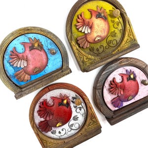 May include: Four decorative, arched wooden plaques with a raised cardinal bird design. Each plaque features a different colour scheme: blue, yellow, red, and pink. The birds are in various shades of red and brown, with a small knob on each plaque.