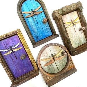 Puede incluir: Cuatro puertas de hadas en miniatura de varias formas y colores. Cada puerta presenta una libélula y un pequeño pomo. Las puertas están pintadas en tonos de azul, morado y marrón, con marcos dorados y de piedra.