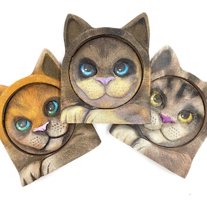 May include: Three cat-shaped coasters with detailed, hand-painted faces. The coasters feature a variety of colours, including brown, orange, and grey, with blue, green, and yellow eyes and pink noses. The coasters are designed to look like cats wearing hoods.